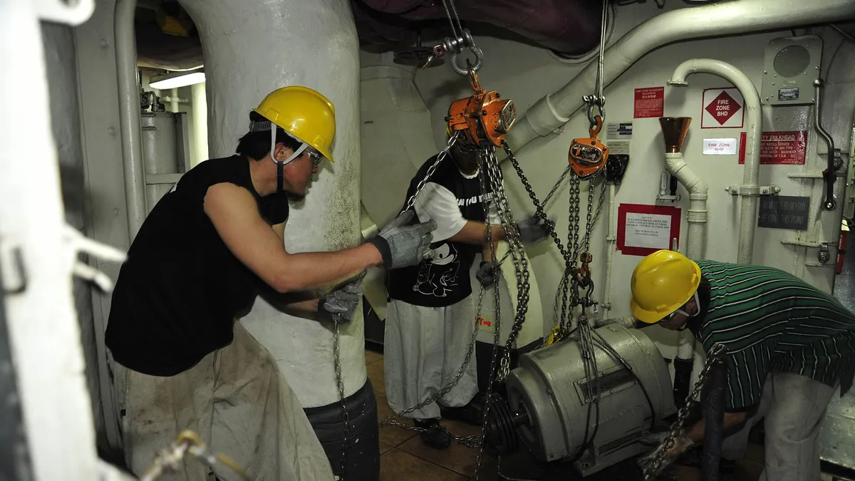 Technicians in hard hats rigging a chain hoist to service machinery in an engine room